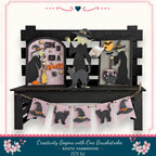 A Halloween-themed display featuring a black wooden bench with various Halloween-themed decorations, including a witch, a cat, and a cauldron. The display also includes a banner with black cat silhouettes and a "One Brushstroke" sign.