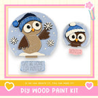 The image is a DIY wood paint kit featuring two wooden owl figures, one larger and one smaller, wearing winter hats and surrounded by snowflakes. The kit includes a sign that says " Owl Be Home for Winter" and a heart symbol, suggesting the kit is designed for creating winter-themed decorations or gifts.