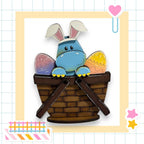 A blue Easter bunny with white ears and a brown basket sits in a basket, surrounded by colorful Easter eggs.
