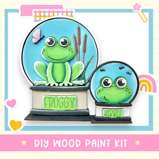 a DIY wood paint kit for creating a frog-themed snow globe. The kit includes a frog figurine and a snow globe, both of which are designed to resemble a frog. The kit is presented in a pink and blue color scheme, with the frog figurine and snow globe being the main focus of the image.