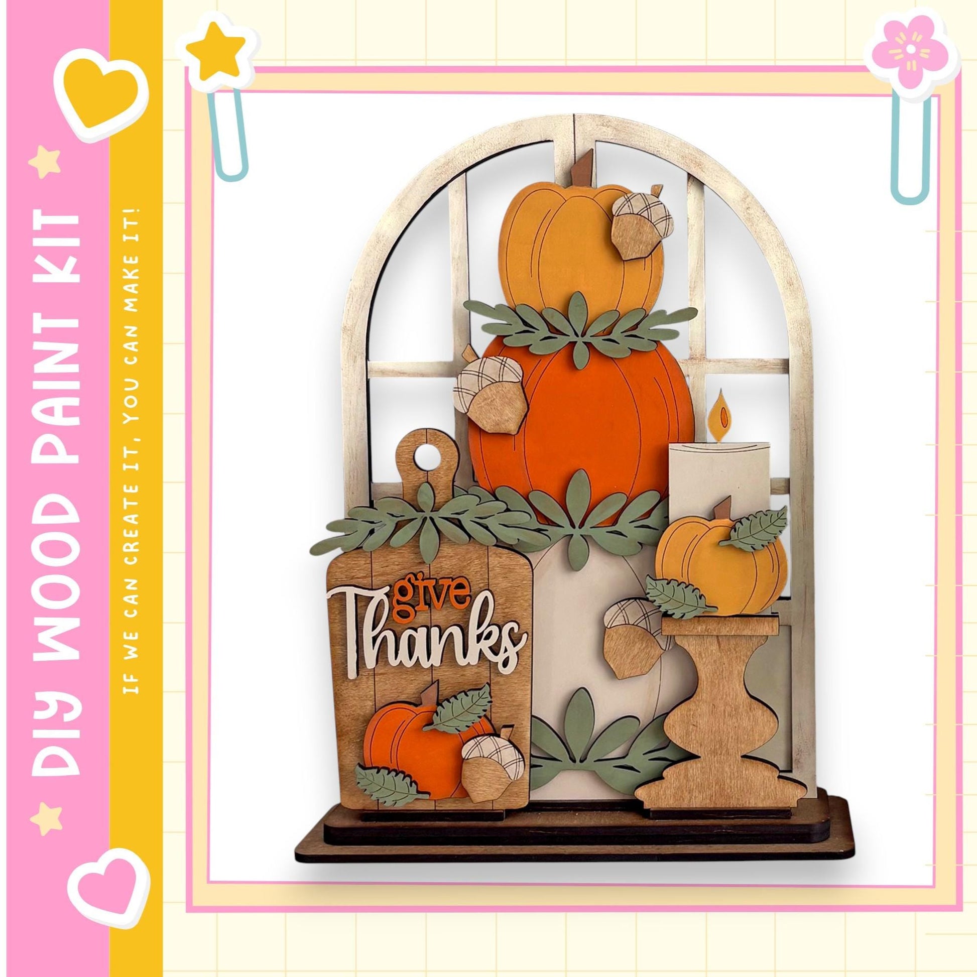A wooden display featuring a carved pumpkin, a candle, and a sign that says "Give Thanks". The display is set against a white background and is framed by a pink border.