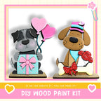 two wooden toy dogs, one holding a gift and the other holding a bouquet of flowers, both wearing hats and bow ties. The image is a collage of two separate images, and the text below the images reads "DIY WOOD PAINT KIT" and "IF WE CAN CREATE IT, YOU CAN MAKE IT!"