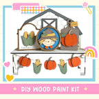 The image is a DIY wood paint kit featuring a scarecrow, pumpkins, and a barn. The kit includes a scarecrow, pumpkins, and a barn, all made of wood and painted in a decorative style.