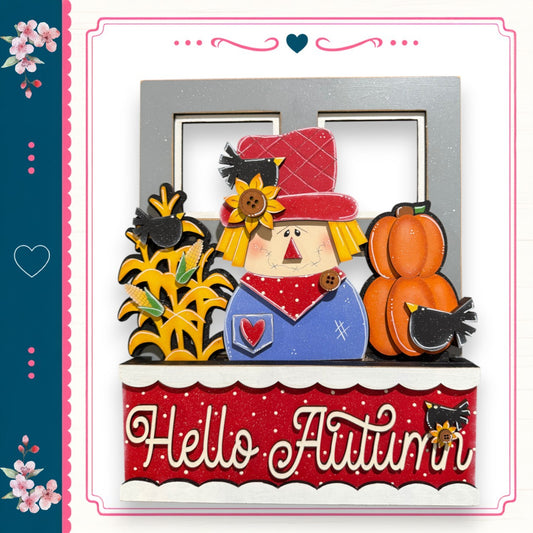 A decorative wooden frame with a scarecrow figure, a pumpkin, and flowers, along with the text "Hello Autumn" displayed on the frame.