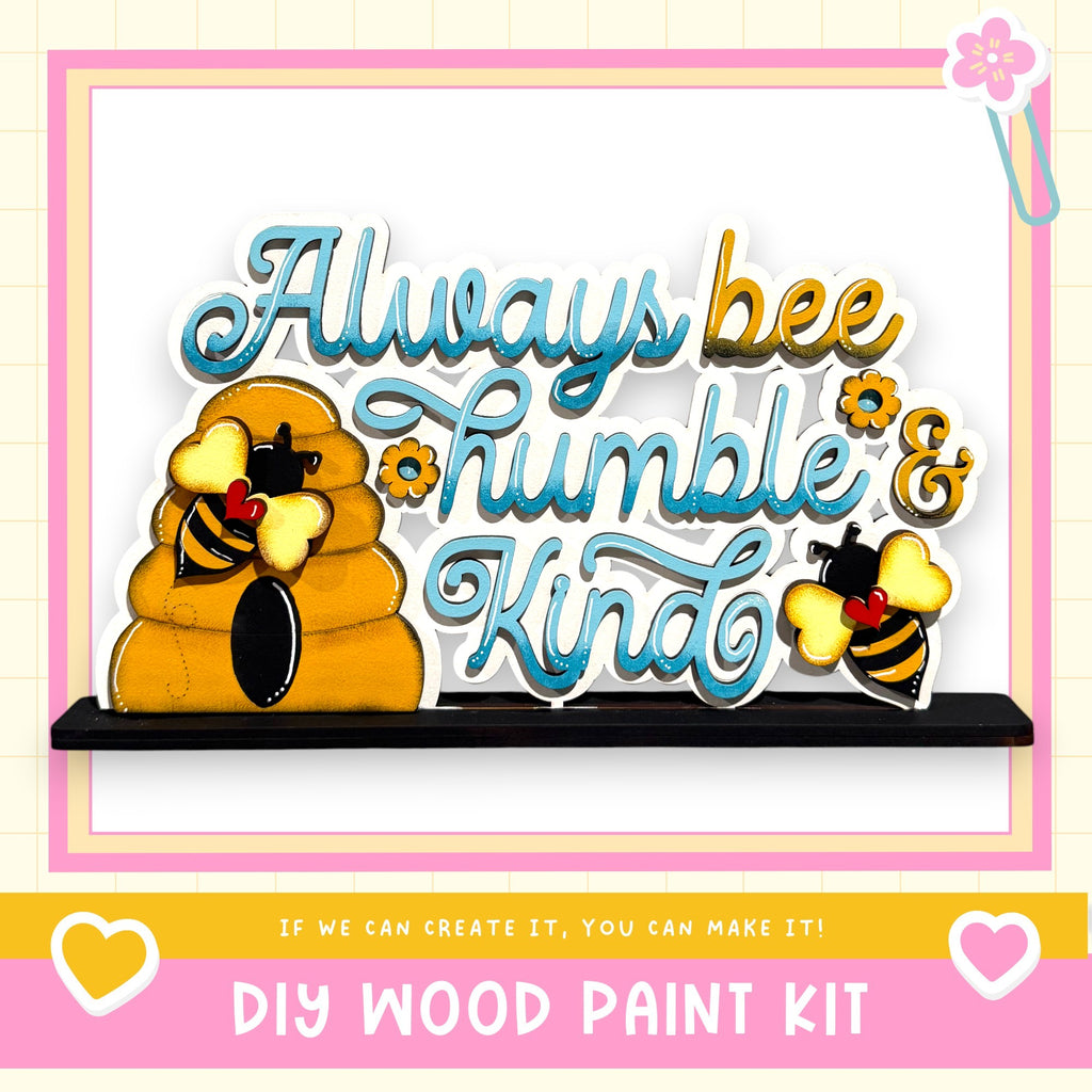 The image is a colorful and playful DIY wood paint kit featuring a beehive and two bees, along with the text "Always bee humbly & kind" and a heart symbol.