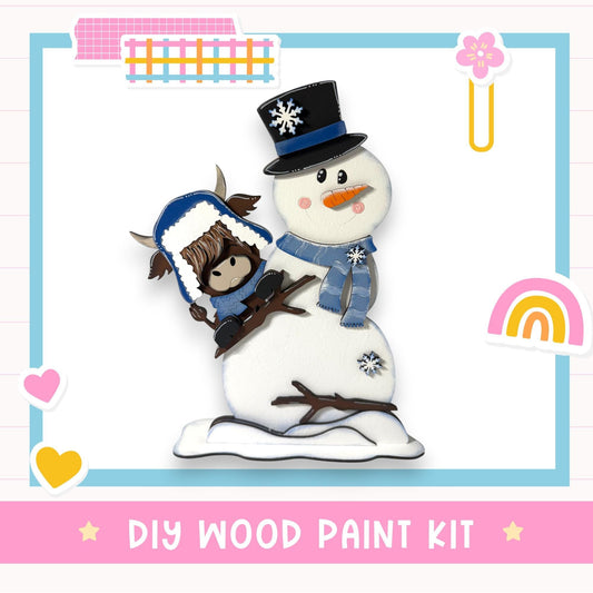 A snowman and a cow figure are depicted in a DIY wood paint kit, surrounded by various colorful stickers and a heart shape.