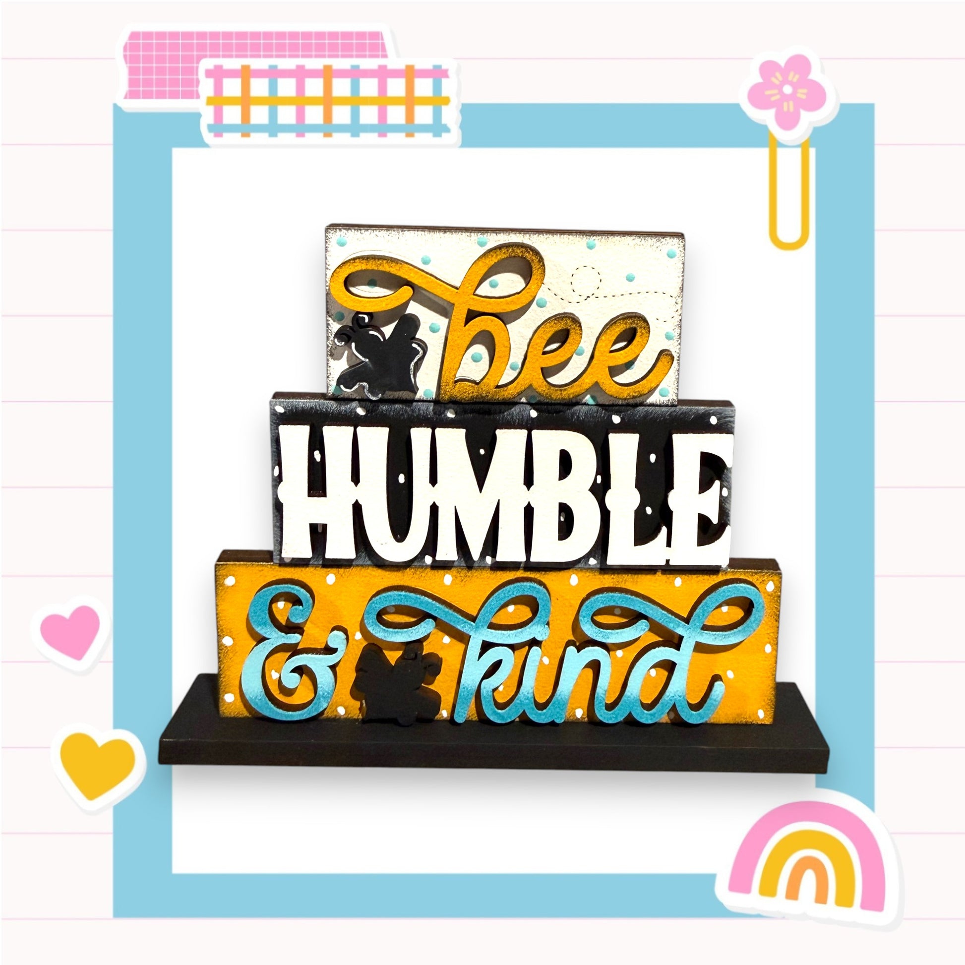 A colorful wooden block with the text "bee humble & kind" written on it, surrounded by various decorative elements such as hearts, flowers, and a rainbow.