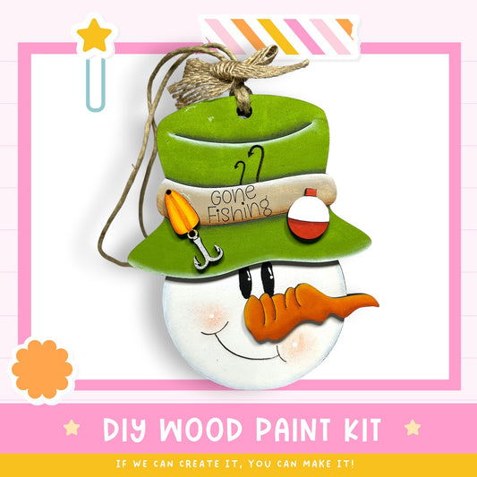 a snowman ornament with a hat, carrot nose, and fishing pole, hanging from a string. The text below the image reads "DIY WOOD PAINT KIT" and "IF WE CAN CREATE IT, YOU CAN MAKE IT!"
