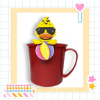 A yellow duck wearing sunglasses is perched on a red mug, set against a grid background with various colorful stickers and a heart-shaped clip.
