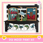 The image is a DIY wood paint kit featuring a wooden bench with a bird, a sign that says "Life is better at the lake", and a banner with various wooden and paper elements.