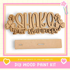 The image is a DIY wood paint kit advertisement, featuring a wooden sign with the text "SHAKE your shamrocks" and a blank wooden board.