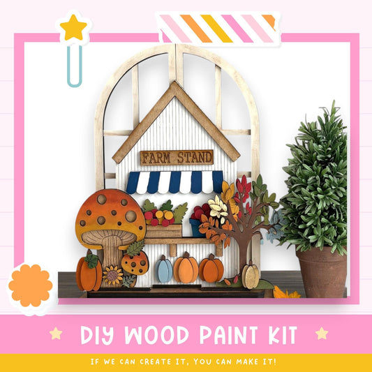 The image is a colorful and whimsical DIY wood paint kit, featuring a farm stand with a mushroom, pumpkins, and a tree, surrounded by a pink and white striped border. The kit appears to be made of wood and is designed for painting, with the text "DIY WOOD PAINT KIT" and "IF WE CAN CREATE IT, YOU CAN MAKE IT!" displayed.