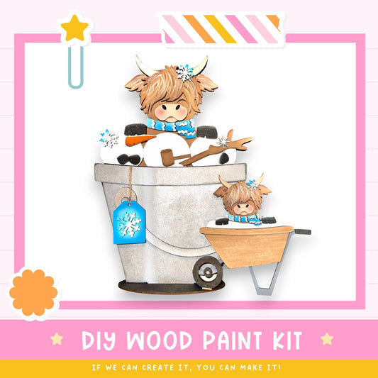 a DIY wood paint kit with a wooden cow figurine sitting in a wheelbarrow, surrounded by snow and other winter-themed elements.