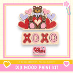 The image is a colorful and playful DIY wood paint kit featuring a teddy bear surrounded by hearts and the text "XOXO" in a playful font.