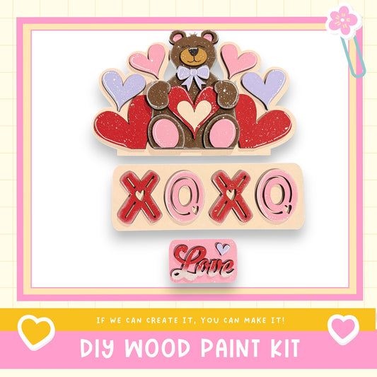 The image is a colorful and playful DIY wood paint kit featuring a teddy bear surrounded by hearts and the text "XOXO" in a playful font.