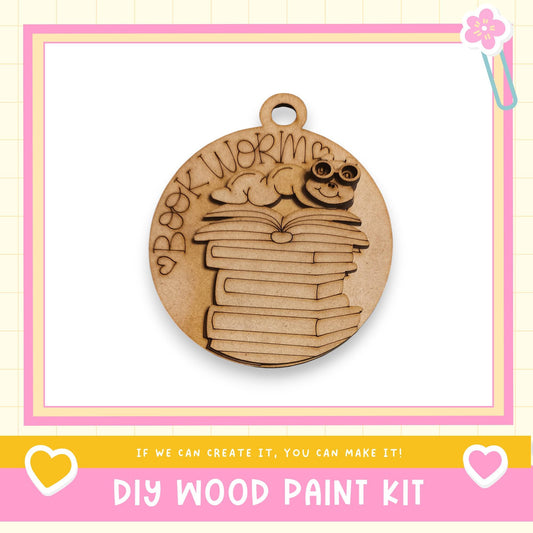 a wooden pendant with a design of stacked books and a smiling character, along with text that says "Book Worm" and "DIY WOOD PAINT KIT".