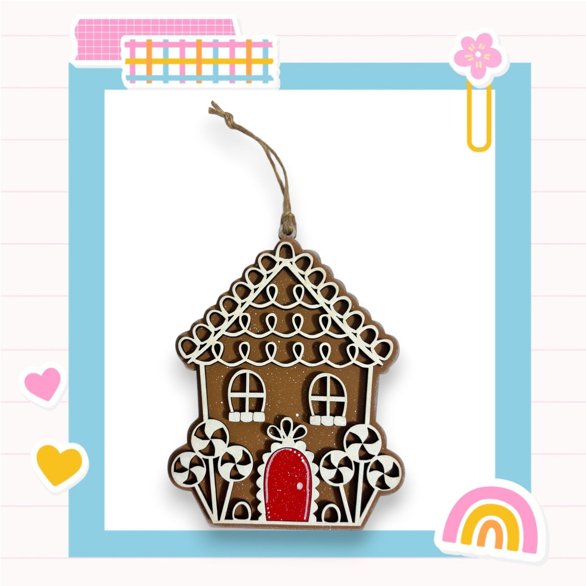 A wooden gingerbread house ornament is displayed against a blue background with various colorful stickers and a pink heart.