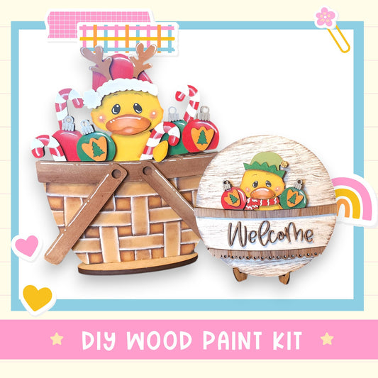 a DIY wood paint kit for creating a wooden basket with a duck and Christmas decorations. The basket is filled with ornaments and a small wooden sign that says "Welcome".