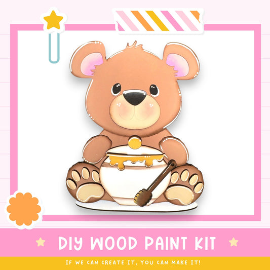 A wooden teddy bear wearing a diaper and holding a spoon, with a pink and white striped border and a star decoration.