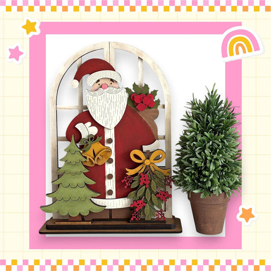 A wooden Santa Claus figure holding a Christmas tree and a potted plant, set against a pink and yellow checkered background with a rainbow and stars.