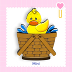 A yellow duck sitting in a brown basket, with a pink heart and a heart-shaped object to the right.
