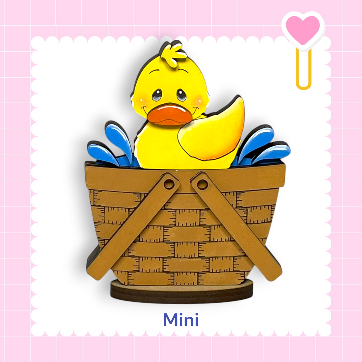 A yellow duck sitting in a brown basket, with a pink heart and a heart-shaped object to the right.