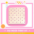The image is a promotional poster for a DIY wood paint kit. It features a heart-shaped stencil with a pattern of pink hearts on a white background, along with a star and a flower. The text on the poster encourages viewers to "If we can create it, you can make it!"