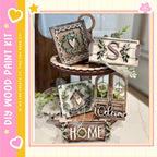 a wooden tray with various decorative items, including a wreath, a sign with the letter "S", and a "Welcome" sign. The tray is placed on a table, and there are additional decorative elements such as leaves and flowers surrounding it.