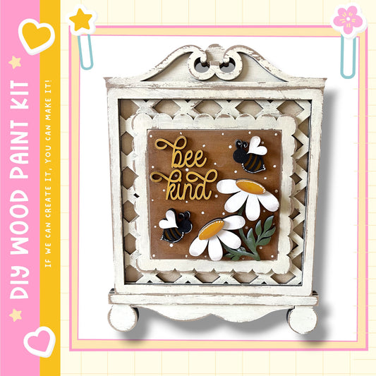 a decorative wooden frame with a floral design and the text "bee kind" written on it. The frame is set against a white background, and there are small flowers and bees on the frame.