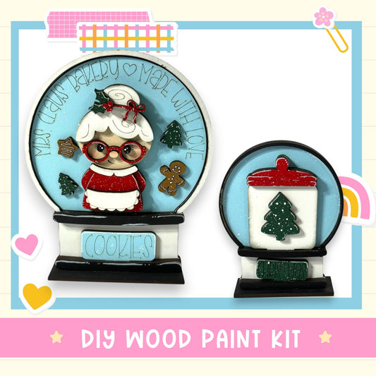 two wooden snowglobe decorations, one with a cookie jar and the other with a Christmas tree. The text "DIY WOOD PAINT KIT" is displayed at the bottom of the image.