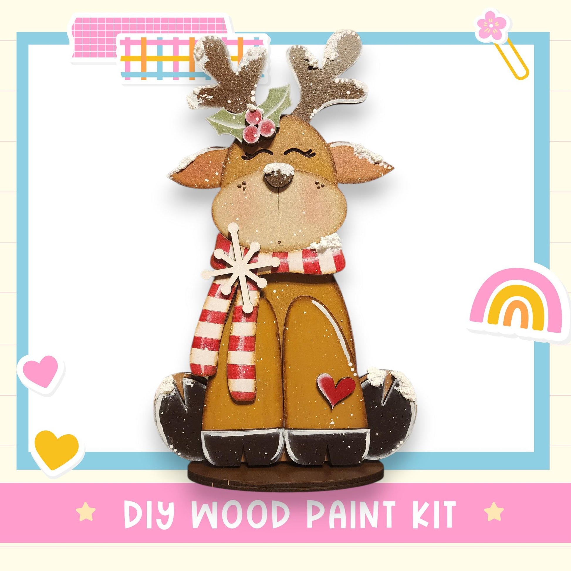 A wooden toy reindeer with antlers and a red heart on its chest, wearing a red and white striped scarf, is the main focus of the image. The background includes various colorful stickers and a rainbow, creating a festive and cheerful atmosphere. The text "DIY WOOD PAINT KIT" is visible, suggesting that this is a DIY wood paint kit for creating a wooden toy reindeer.
