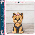 A wooden cutout of a Yorkshire Terrier dog with a pink bow on its head, sitting on a wooden surface against a white background.