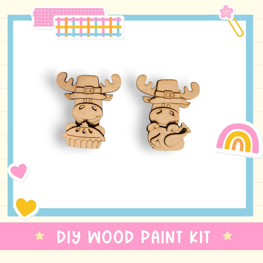 two wooden animal figurines, likely moose or deer, wearing festive hats and holding festive items such as a cake or cupcake. The image also includes various colorful stickers and a rainbow-colored heart, suggesting a festive or holiday theme. The text "DIY WOOD PAINT KIT" is visible, indicating that this is a DIY wood paint kit for creating wooden decorations or toys.