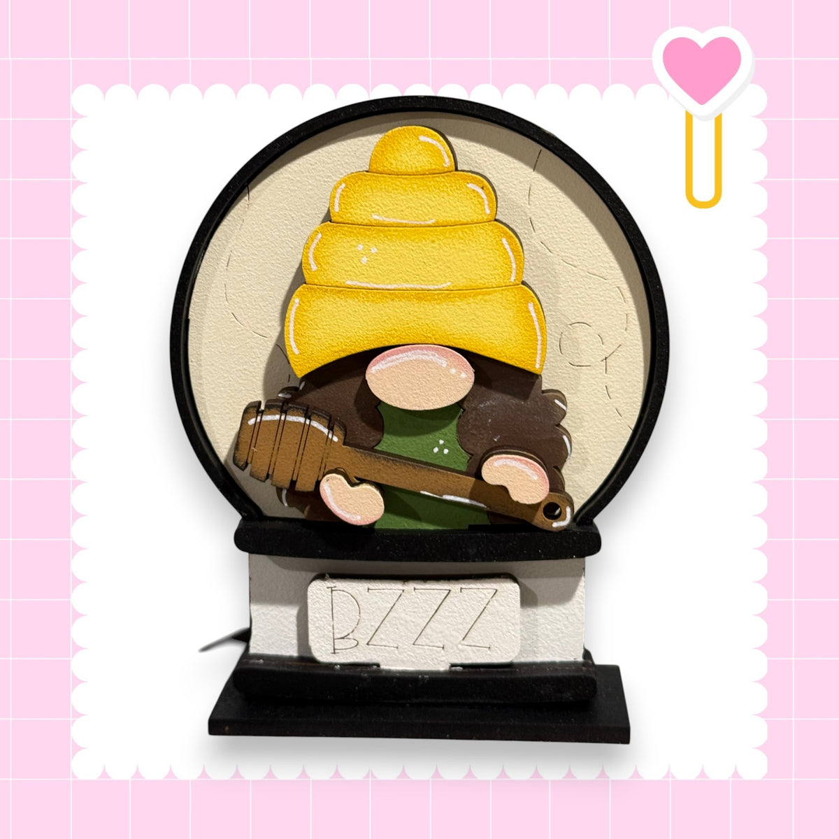 A gnome wearing a yellow hat and holding a guitar is depicted in the center of the image, set against a pink background with a heart shape and a small yellow heart shape.