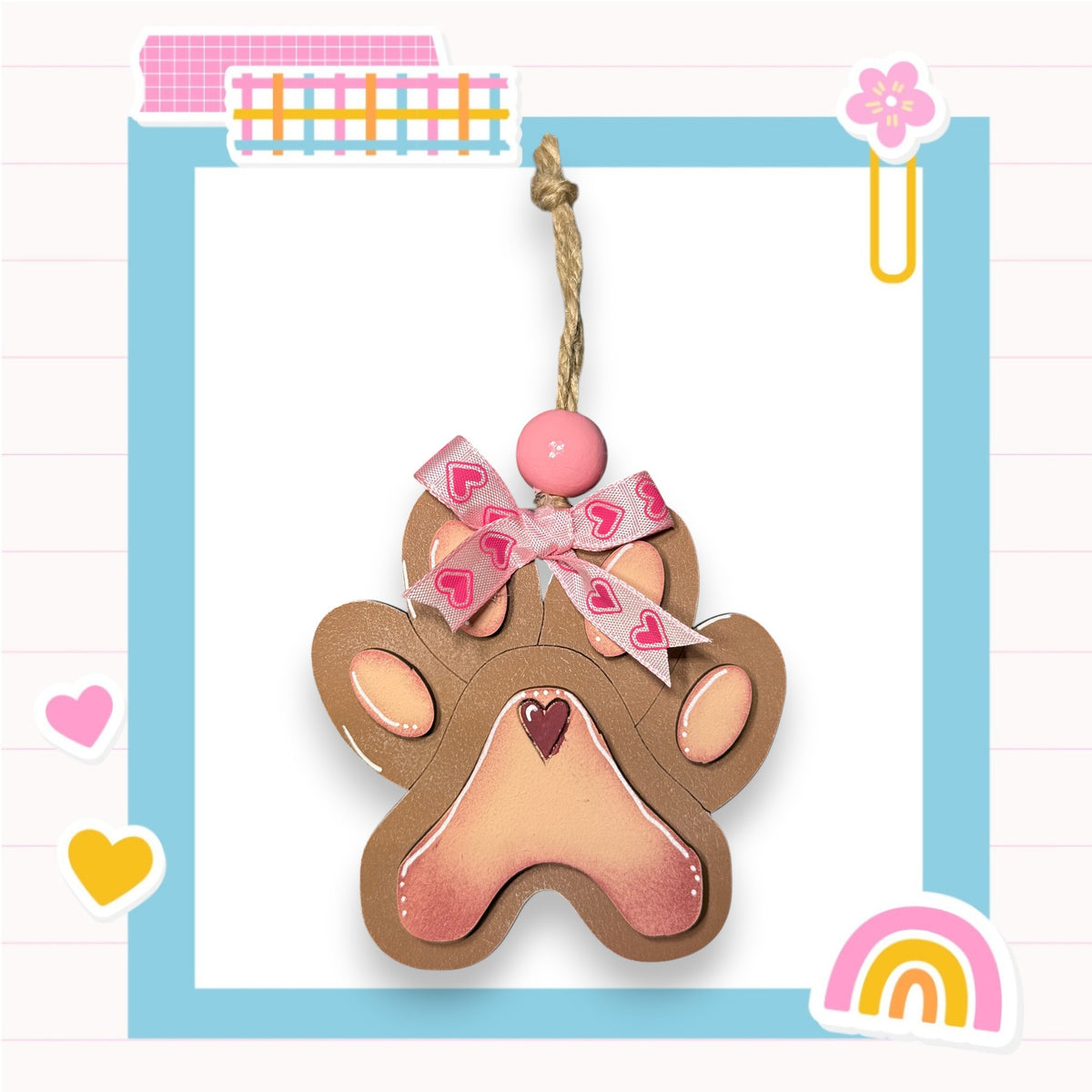 A brown paw print-shaped ornament with a pink ribbon and heart-shaped embellishments, hanging from a string.