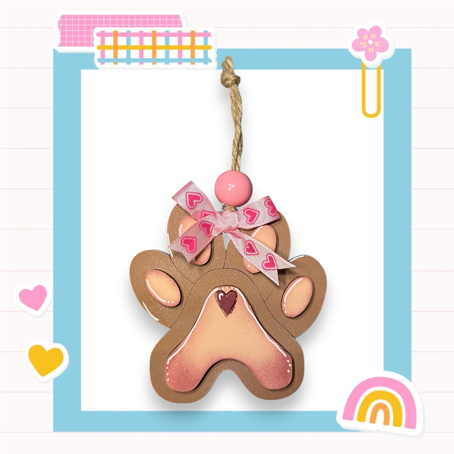A brown paw print-shaped ornament with a pink ribbon and heart-shaped embellishments, hanging from a string.