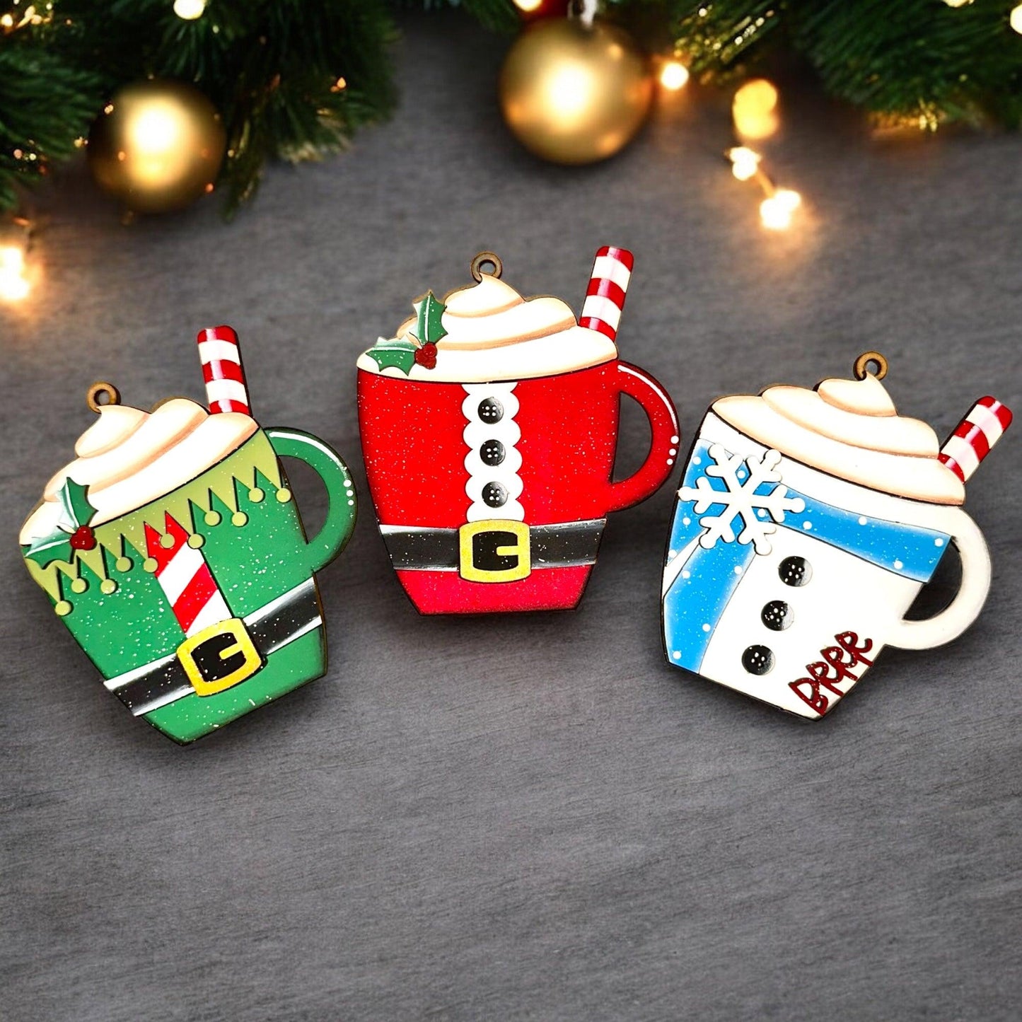 three christmas themed coffee mugs sitting next to a christmas tree