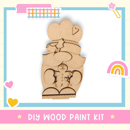 a DIY wood paint kit, including a wooden gnome and a heart-shaped object, set against a light blue background with a pink border.
