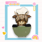 A cartoon cow wearing a chef's hat is sitting in a green bowl, surrounded by various colorful and whimsical elements such as hearts, a rainbow, and a flower.