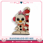 A hand-made wooden snowman figure with a red and white striped pole, wearing a pink bow and earmuffs, sitting on a white base.