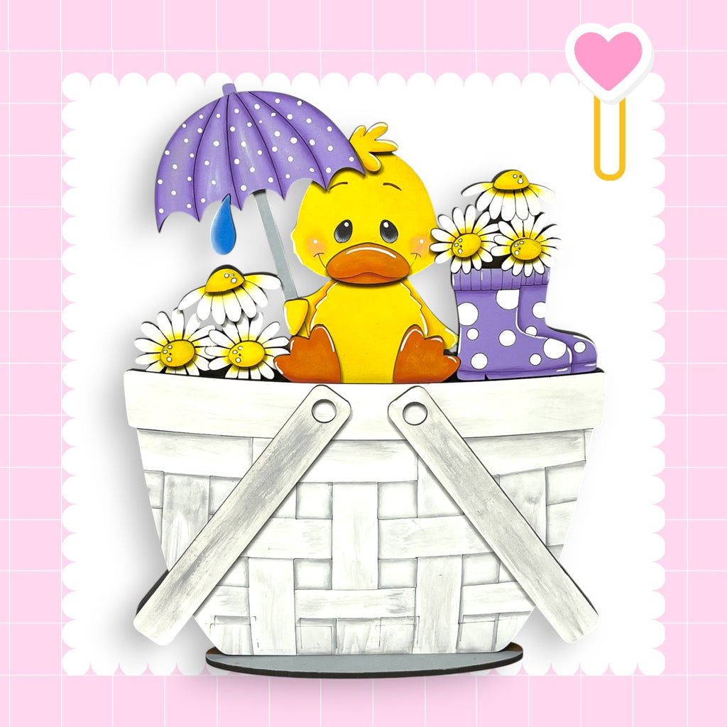 A yellow duck sitting in a white basket filled with flowers and an umbrella, set against a pink background with a heart shape.