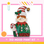 a DIY wood paint kit for creating a wooden elf decoration. The kit includes a wooden elf figure, a candy cane, and the word "Believe" written on the elf's chest. The kit is presented in a pink and white color scheme, with a star decoration on the left side. The text "DIY WOOD PAINT KIT" is displayed at the bottom of the image, along with the phrase "IF WE CAN CREATE IT, YOU CAN MAKE IT!"