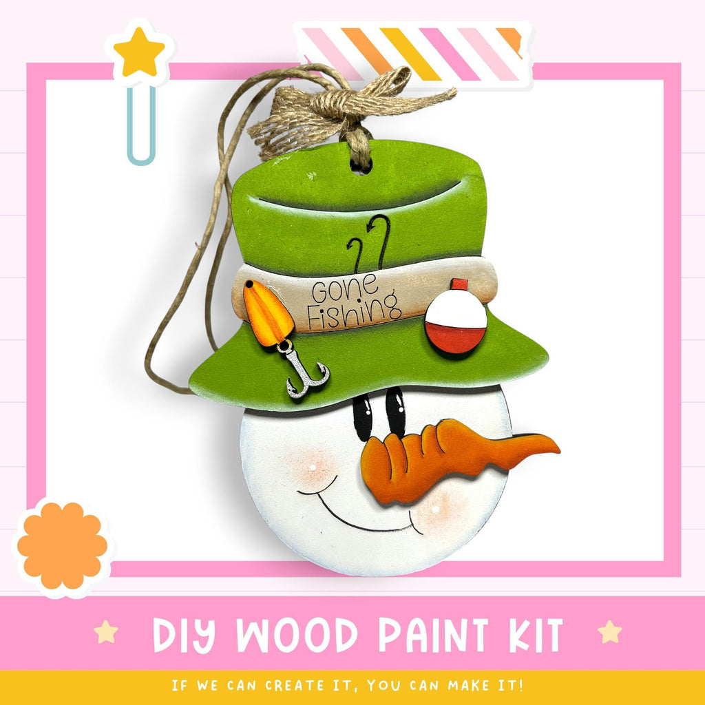 a snowman ornament with a hat, carrot nose, and fishing pole, hanging from a string. The text below the image reads "DIY WOOD PAINT KIT" and "IF WE CAN CREATE IT, YOU CAN MAKE IT!"