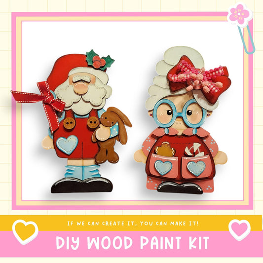 two wooden figures, Santa Claus and Grandma, with Santa holding a teddy bear and Grandma wearing a red bow. The image is framed with a pink border and includes text that reads "DIY WOOD PAINT KIT" and "If we can create it, you can make it!"