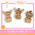 three wooden animal figurines wearing hats, likely representing moose or deer, set against a pink and white background.