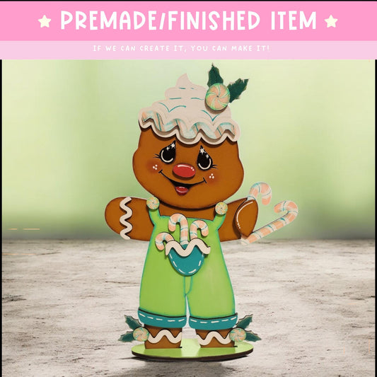 A gingerbread man figure wearing a green outfit and holding a candy cane, with a wreath on its head, standing on a green base.