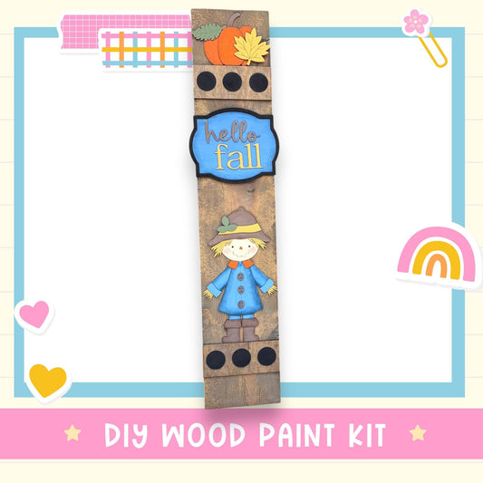 The image is a colorful and playful DIY wood paint kit featuring a wooden stick with a cartoon character and fall-themed decorations.