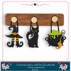 A DIY kit featuring a wooden board with three Halloween-themed charms hanging from it, including a witch's hat, a black cat, and a cauldron with a green substance.