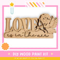 The image is a promotional poster for a DIY wood paint kit. It features a wooden sign with the word "LOVE" written on it, along with a cute cartoon character resembling a cherub or angel. The poster emphasizes that the kit allows for creativity and encourages viewers to create their own designs.