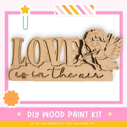 The image is a promotional poster for a DIY wood paint kit. It features a wooden sign with the word "LOVE" written on it, along with a cute cartoon character resembling a cherub or angel. The poster emphasizes that the kit allows for creativity and encourages viewers to create their own designs.